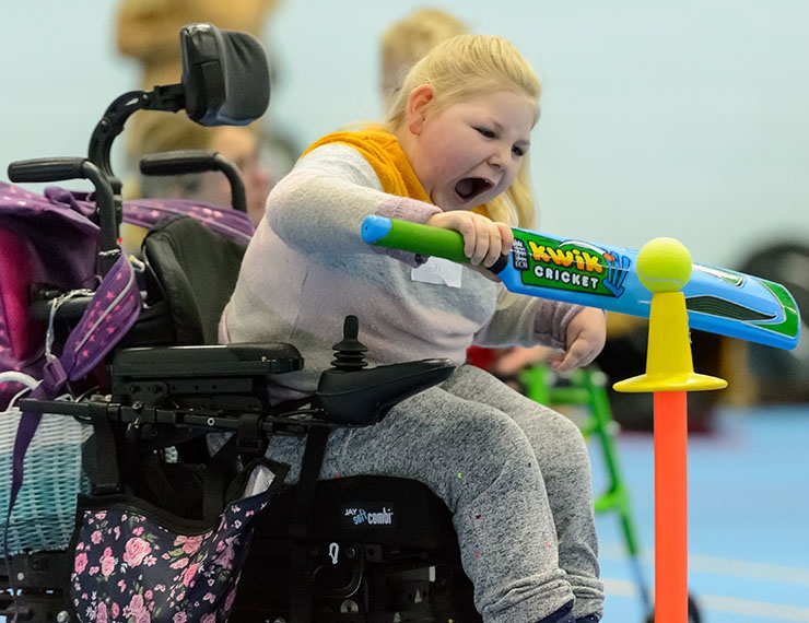 A girl with cerebral palsy, in a wheelchair, hits a ball with a cricket bat, from a tee