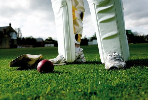 A cricket bat and ball on the grass with a cricketer's legs in the background