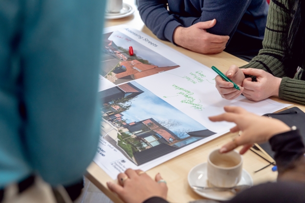 An overhead photo of a table during a planning workshop, on which are people's hands and an annotated diagram of a development