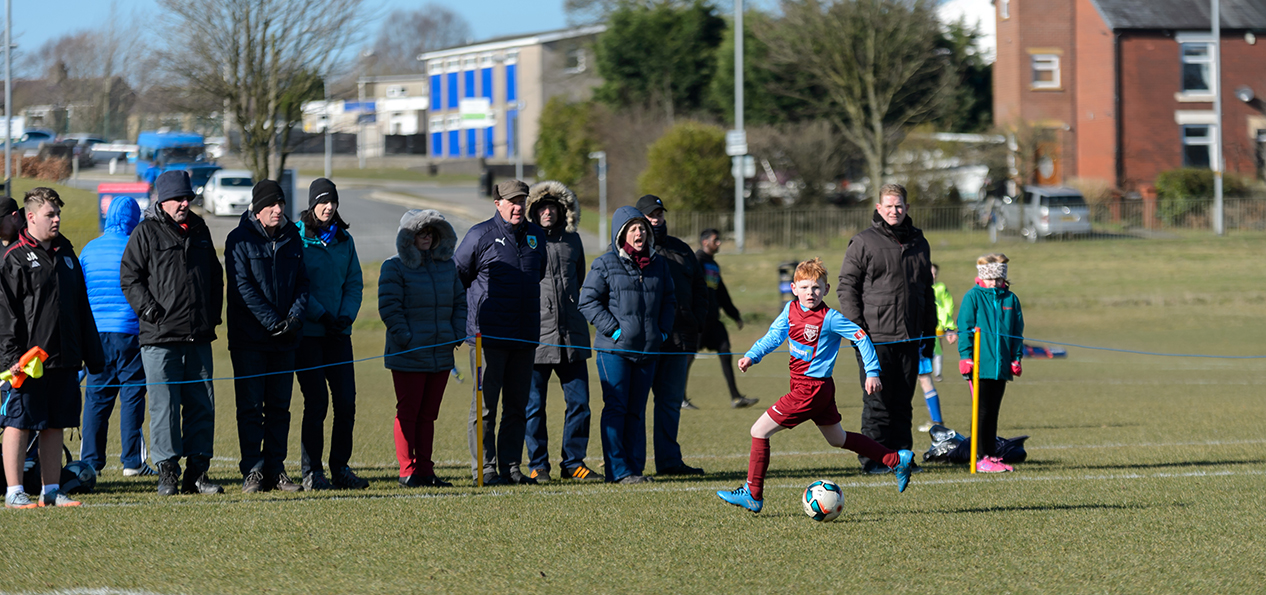 A group of people watching a boy's football match on a playing field.