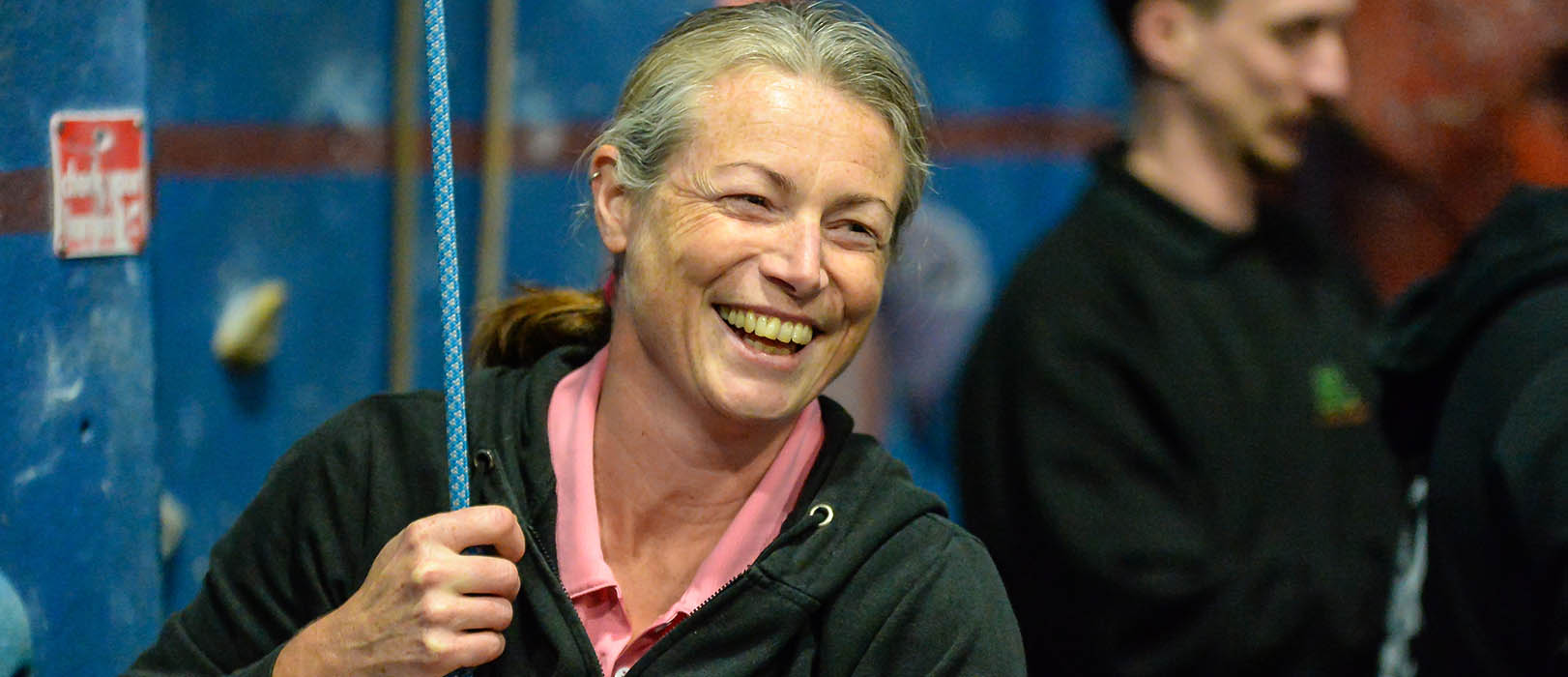 A woman smiles while holding a rope at the foot of an indoor climbing wall