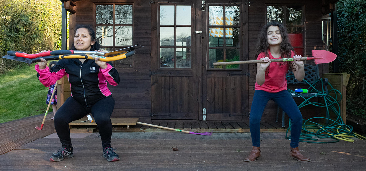 A mother and daughter exercise in their garden, using garden tools as weights