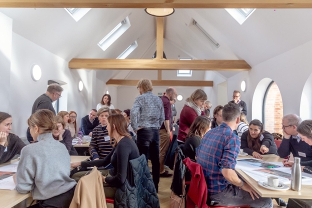 A group of people with documents sit and talk at tables in a hall with exposed beams during a workshop.