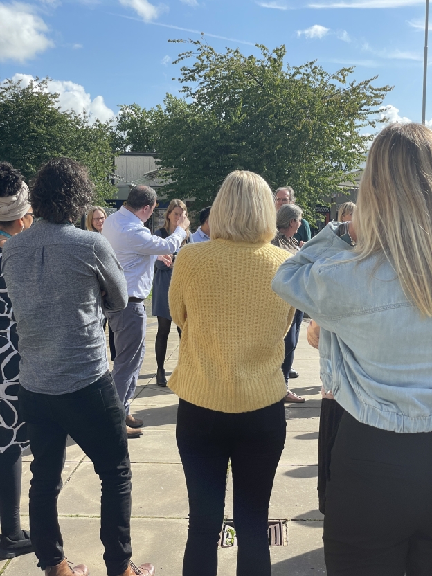 A man gesticulates while talking to a group of people circled around him outdoors.