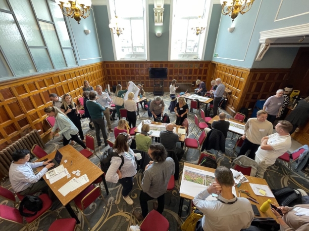 A group of people sit with documents at tables in a hall during a workshop.