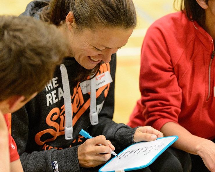 Three people sit on a bench in a sports hall, writing on a mini white board