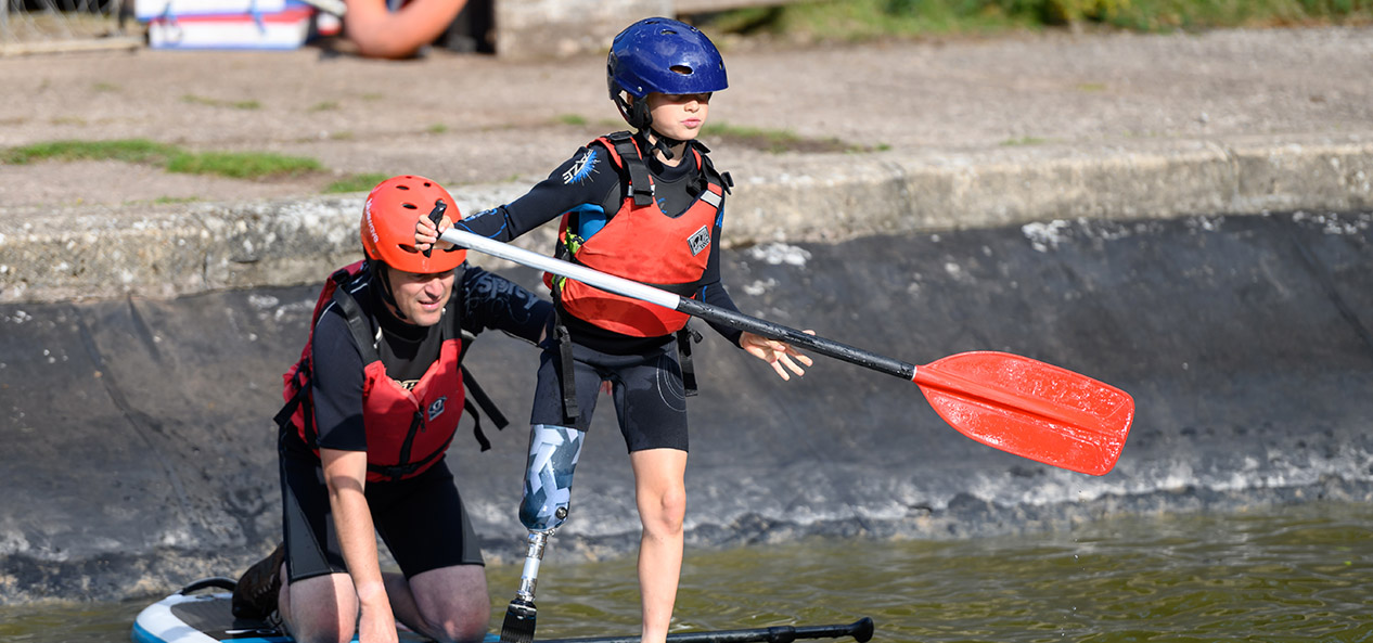 A boy with a prosthetic leg paddles a paddle board, with an adult assistant on the back of it.