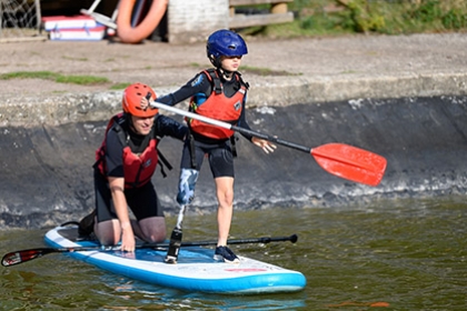 A boy with a prosthetic leg paddles a paddle board, with an adult assistant on the back of it.