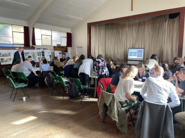 A group of people sit with documents at tables in a hall during a workshop.