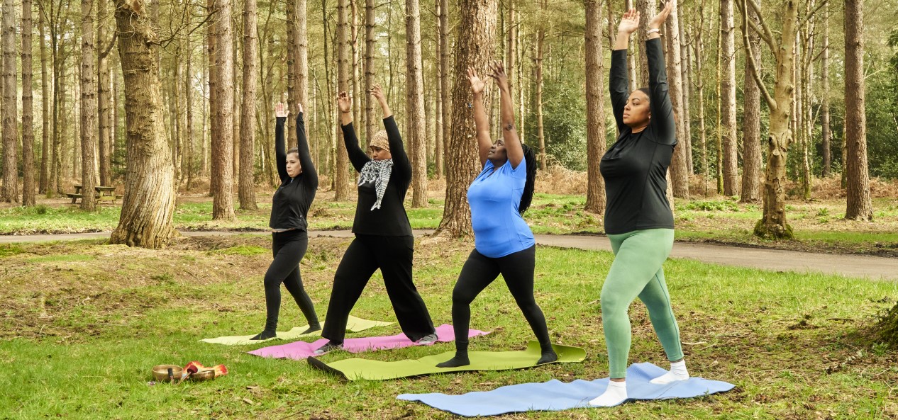 Four women stretch upwards while stood side-by-side on towels in a woodland area.