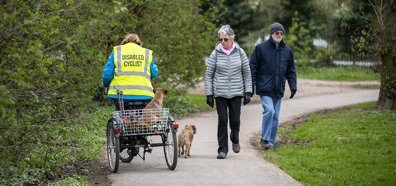 A disabled cyclist, riding a trike, rides with her down past an elderly couple on a path.