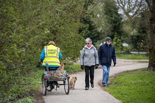 A disabled cyclist, riding a trike, rides with her down past an elderly couple on a path.
