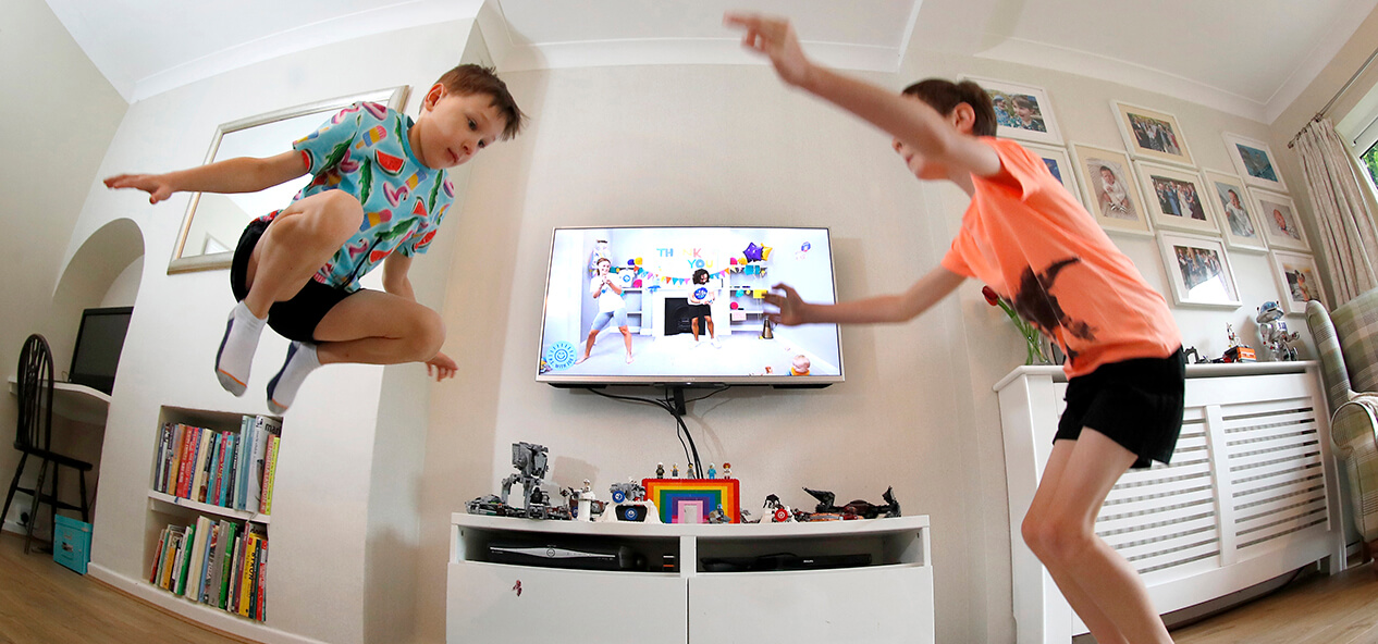 Two boys jumping as they follow along with a Joe Wicks workout playing on the TV behind them.