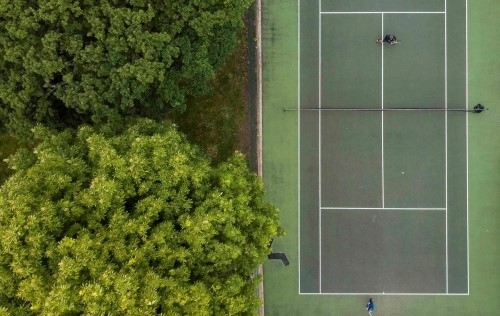 An overhead shot of a green tennis court in a park, next to green trees.