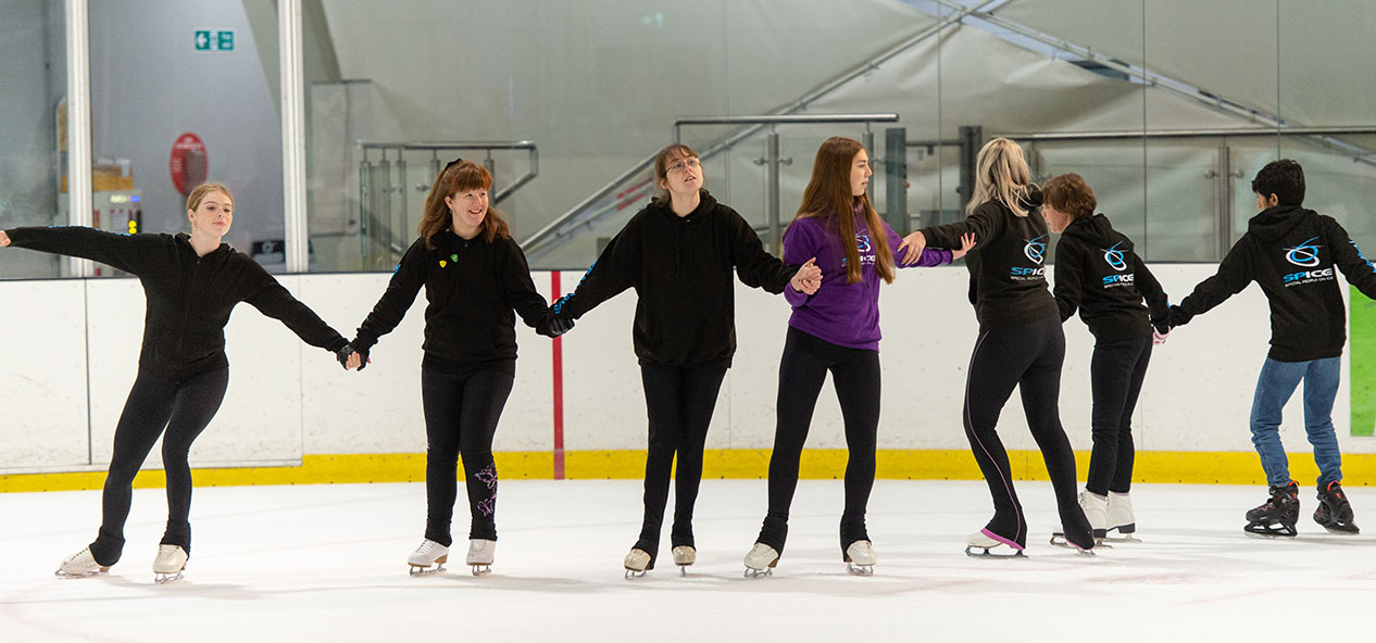 Some young girls and a boy skate holding each other arms and making a big circle on an indoors ice rink.