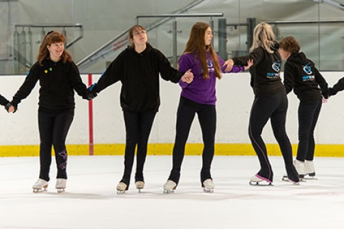 Some young girls and boy skate holding each other arms and making a big circle on an indoors ice rink.