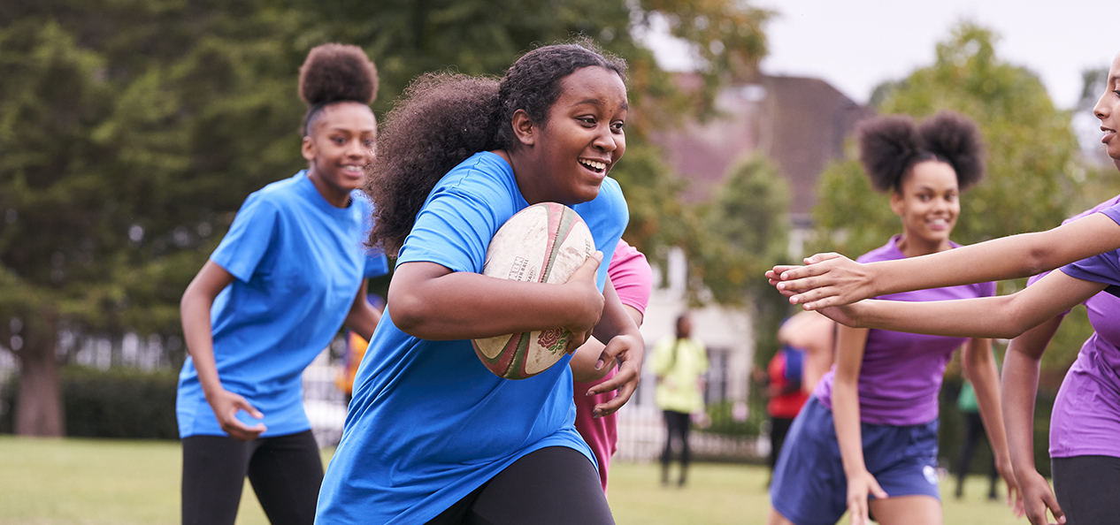 A girl runs with a rugby ball as a group of girls play rugby in a park.