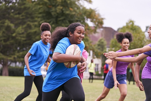 A girl runs with a rugby ball as a group of girls play rugby in a park.