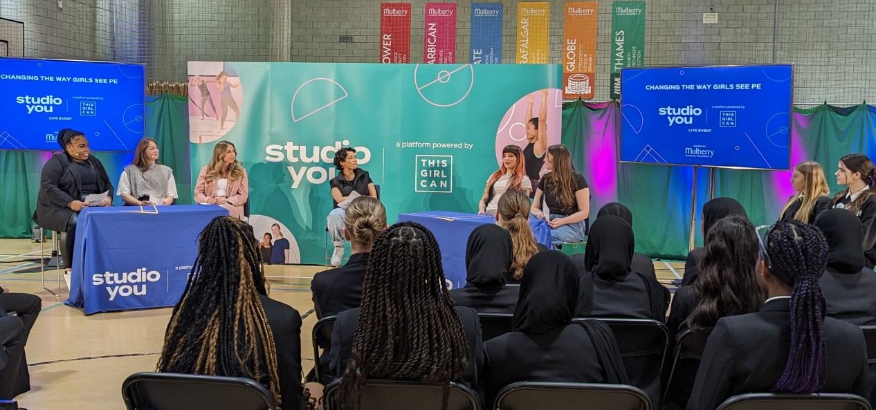 A group of secondary school students sat on chairs in a sports hall watch a panel discussion between a host and five women.