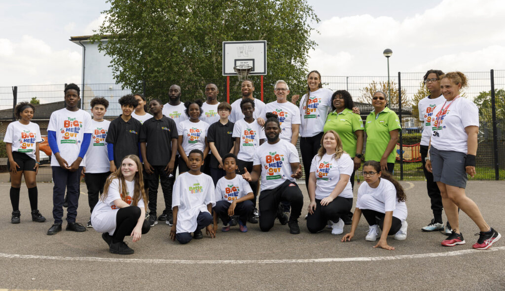 A group of adults and young people pose for a picture on an outdoors basketball pitch with most of them wearing white T-shirts that have The Big Help Out logo on them