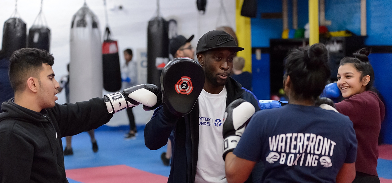 Three young people in a boxing gym gather round their coach as he demonstrates using boxing pads