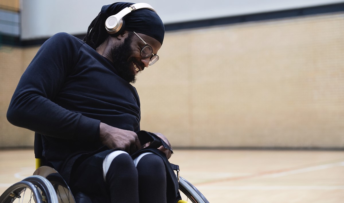 A man wearing headphones smiles while playing wheelchair basketball in a sports hall