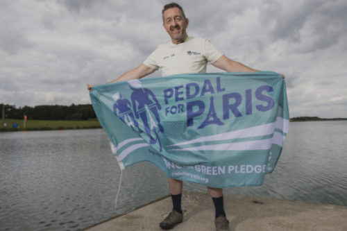 Chris Boardman holds a Pedal for Paris flag in front of a lake