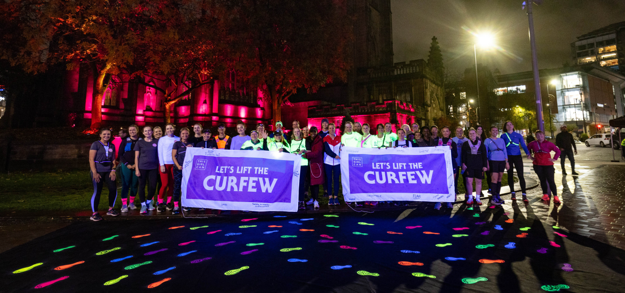 Runners pose with Let's Lift the Curfew signs in front of glow-in-the-dark footprints at night in Manchester